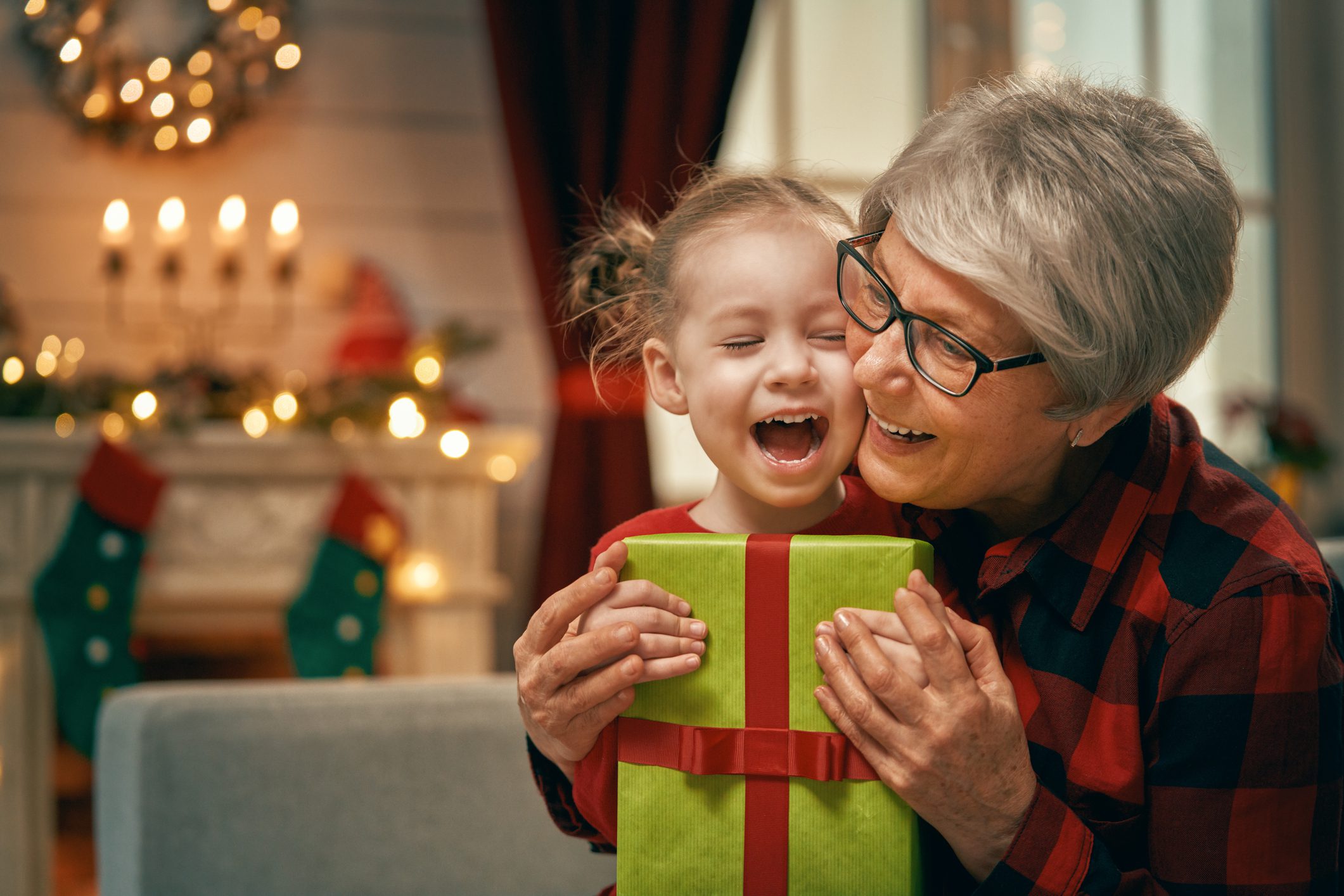 A grandmother hugs her granddaughter who is holding a holiday present. The room is decorated festively for the holidays.