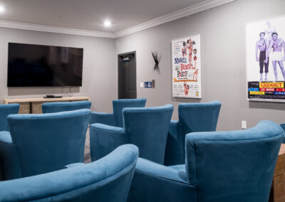 The theater room at Prime Carolina Coast, featuring a large flat-screen TV, plush blue armchairs, and vintage movie posters.